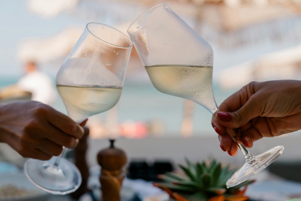 Current image: Two people toasting with white wine glasses at a beach setting. A perfect moment of celebration and relaxation by the sea.